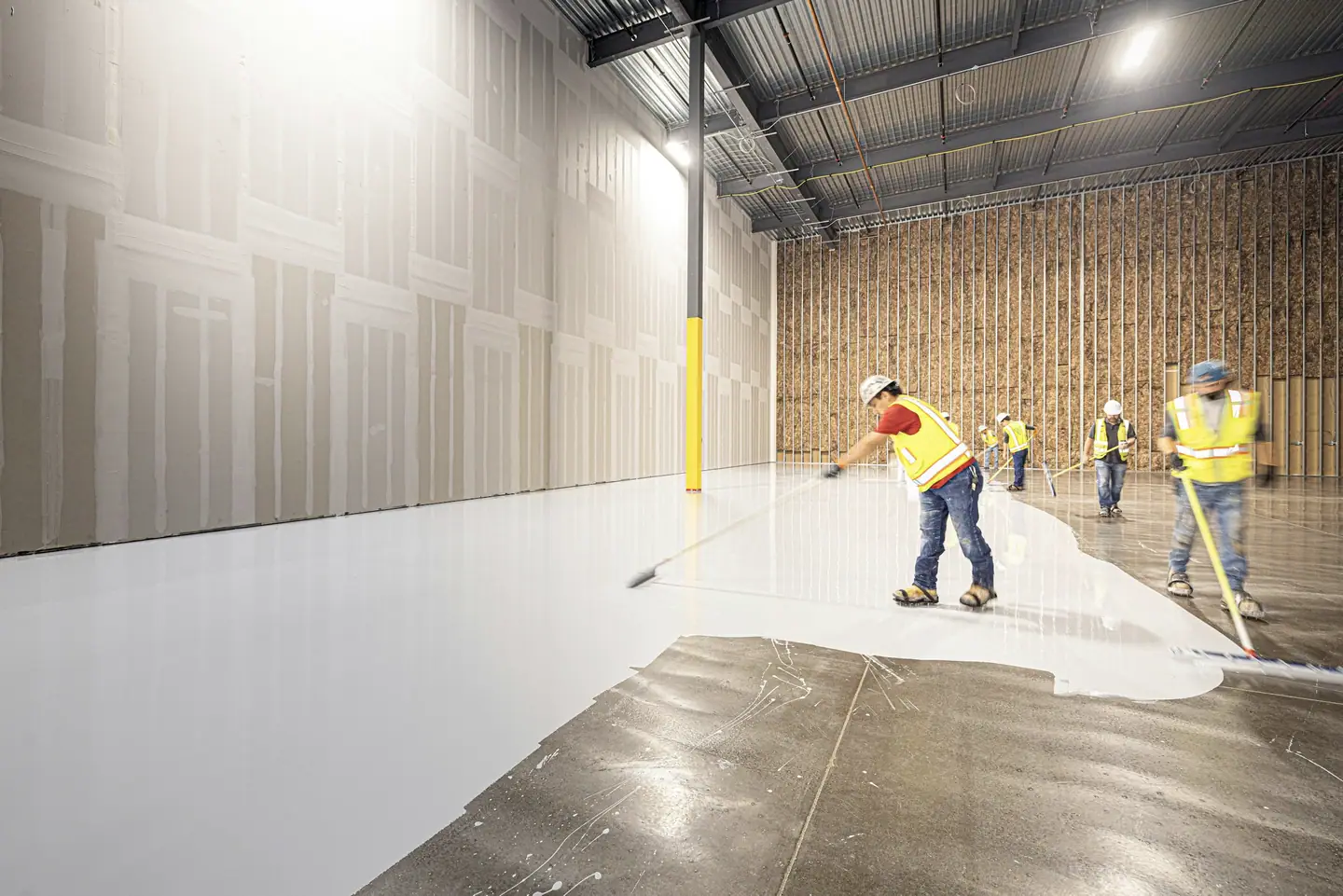 Crew members in hard hats and safety vests applying a white floor coating across a large commercial building interior