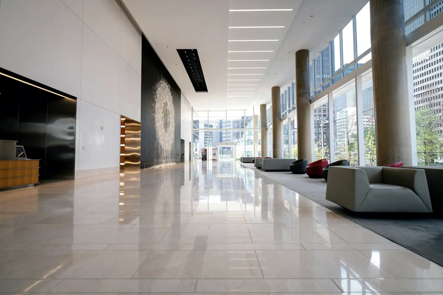 Bright corporate office lobby with polished stone floors reflecting daylight from a floor-to-ceiling glass wall, lounge chairs lined along the windows