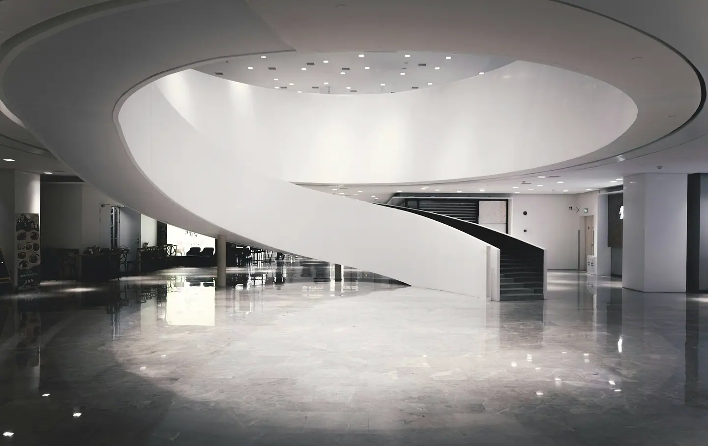 Sculptural white spiral staircase rising through an oval opening in a building lobby with a glossy stone floor